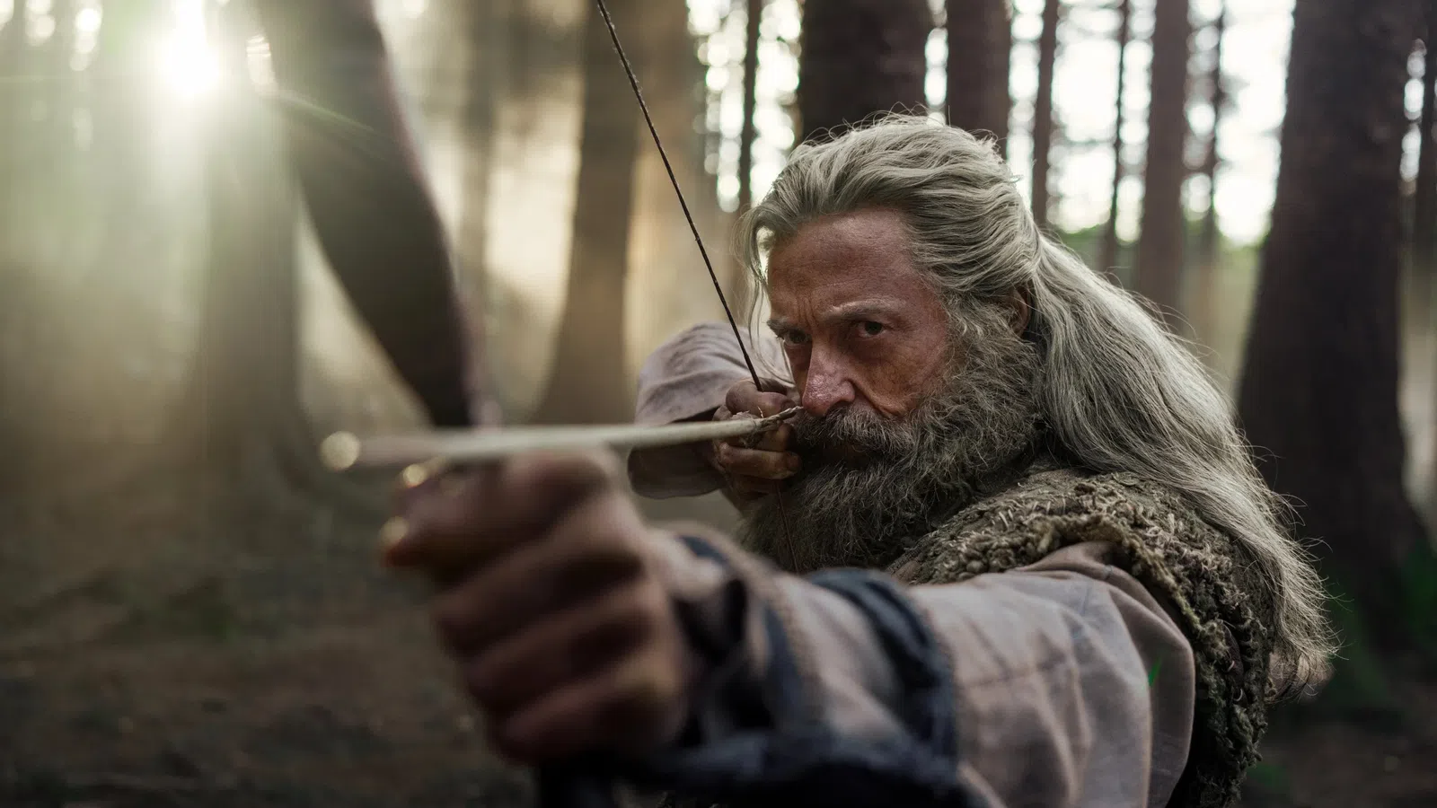 Focused bearded man with long hair aiming a bow and arrow in a misty forest during daytime