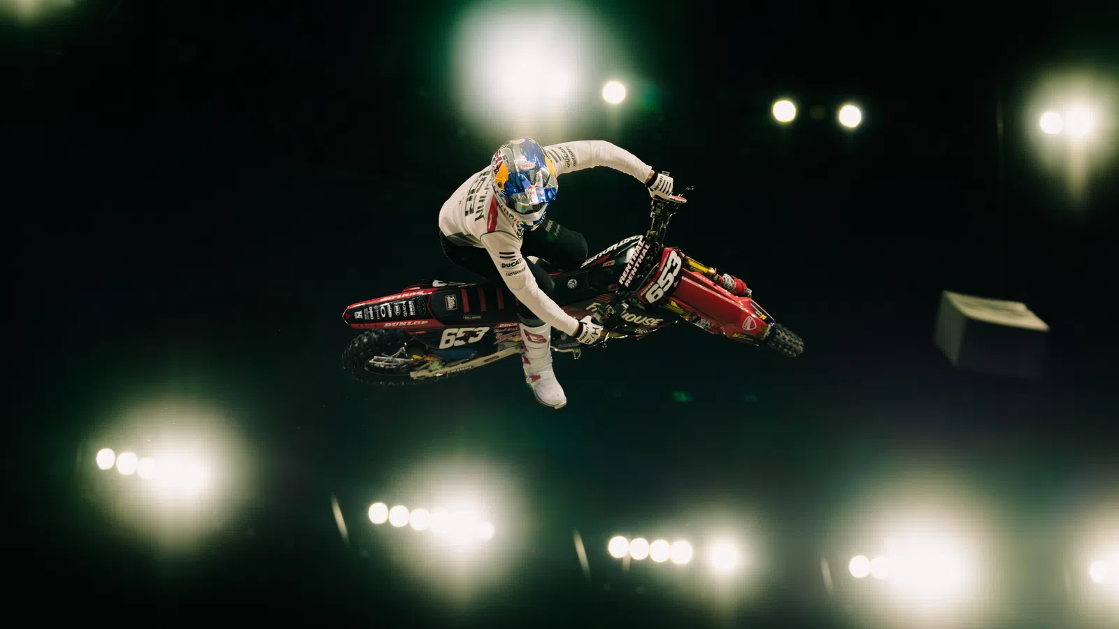 Motocross rider performing a stunt mid-air against bright stadium lights in a dark arena.