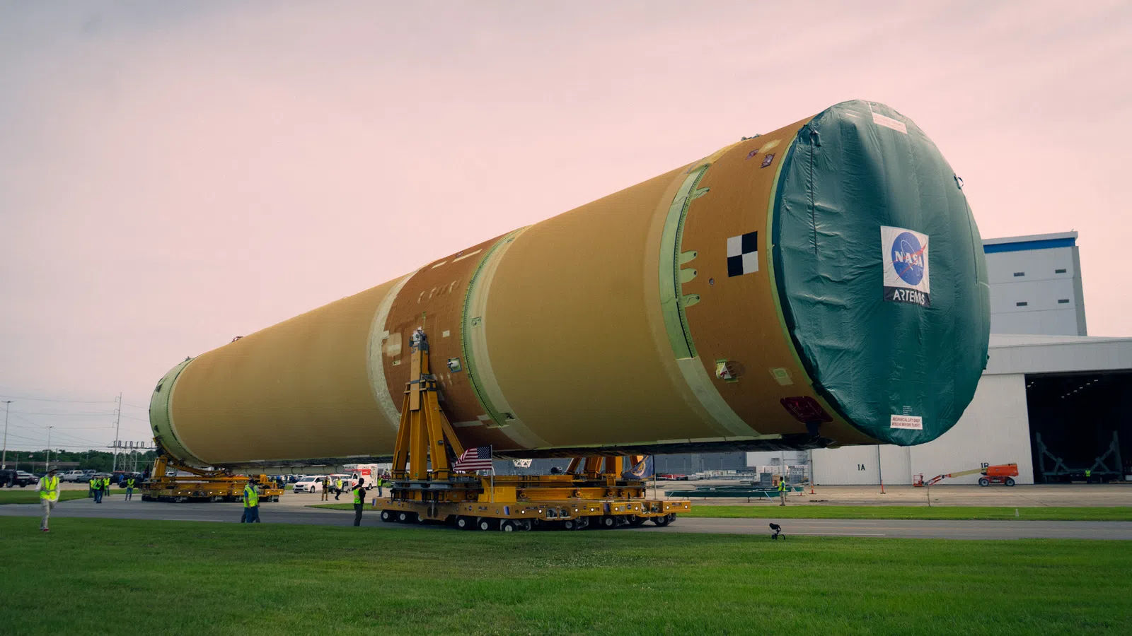 Large NASA Artemis rocket stage being transported on wheeled platforms outside a space facility at dusk.