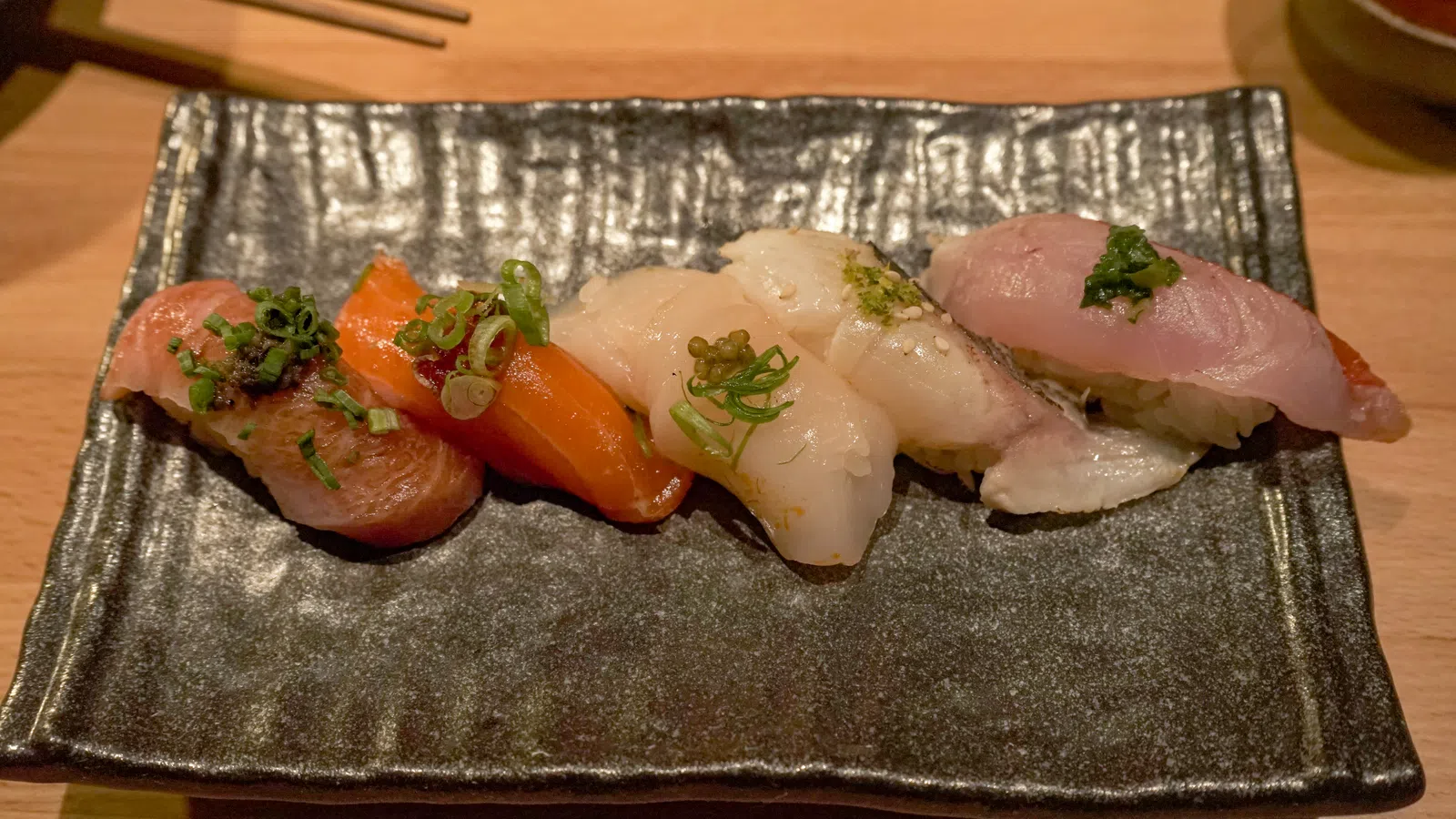 Assorted nigiri sushi with salmon, tuna, scallop, and white fish served on a black rectangular plate.