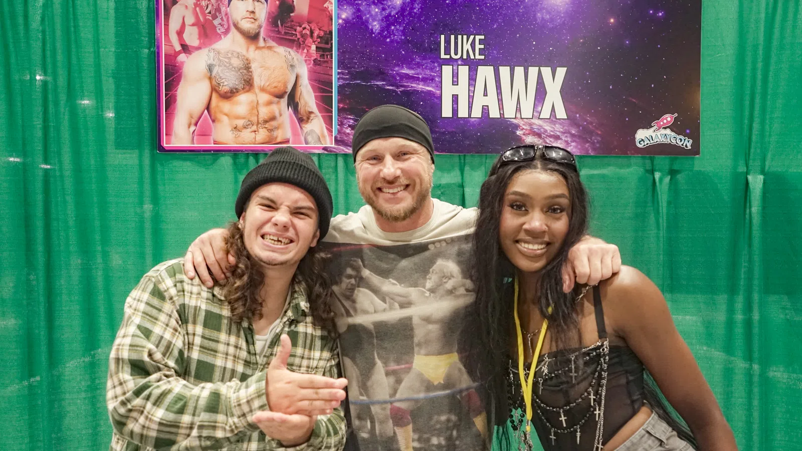 Three people smiling and posing in front of a banner featuring wrestler Luke Hawx at a convention booth.