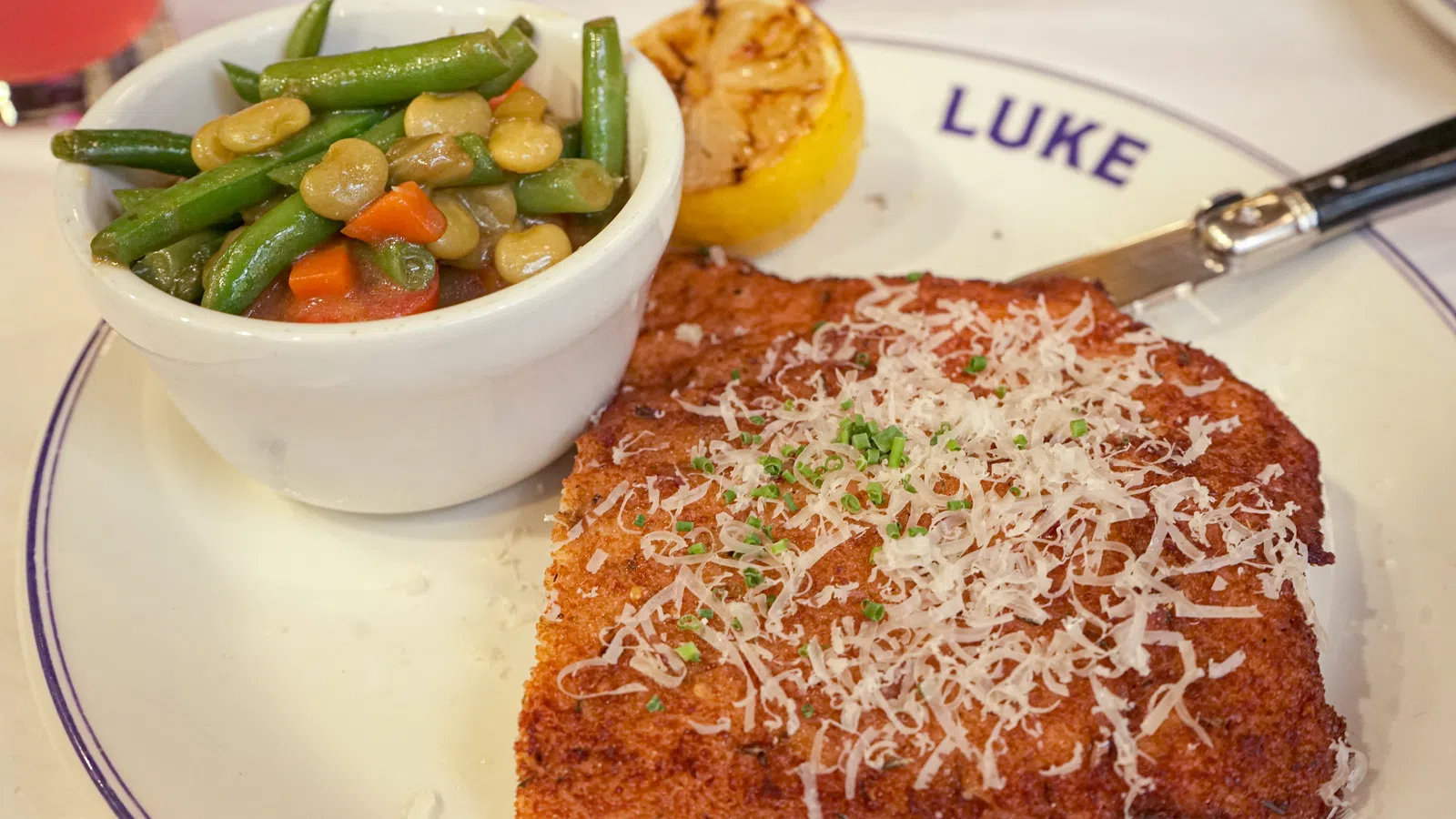Breaded meat cutlet topped with grated cheese and herbs served with mixed vegetables and grilled lemon on a white plate.