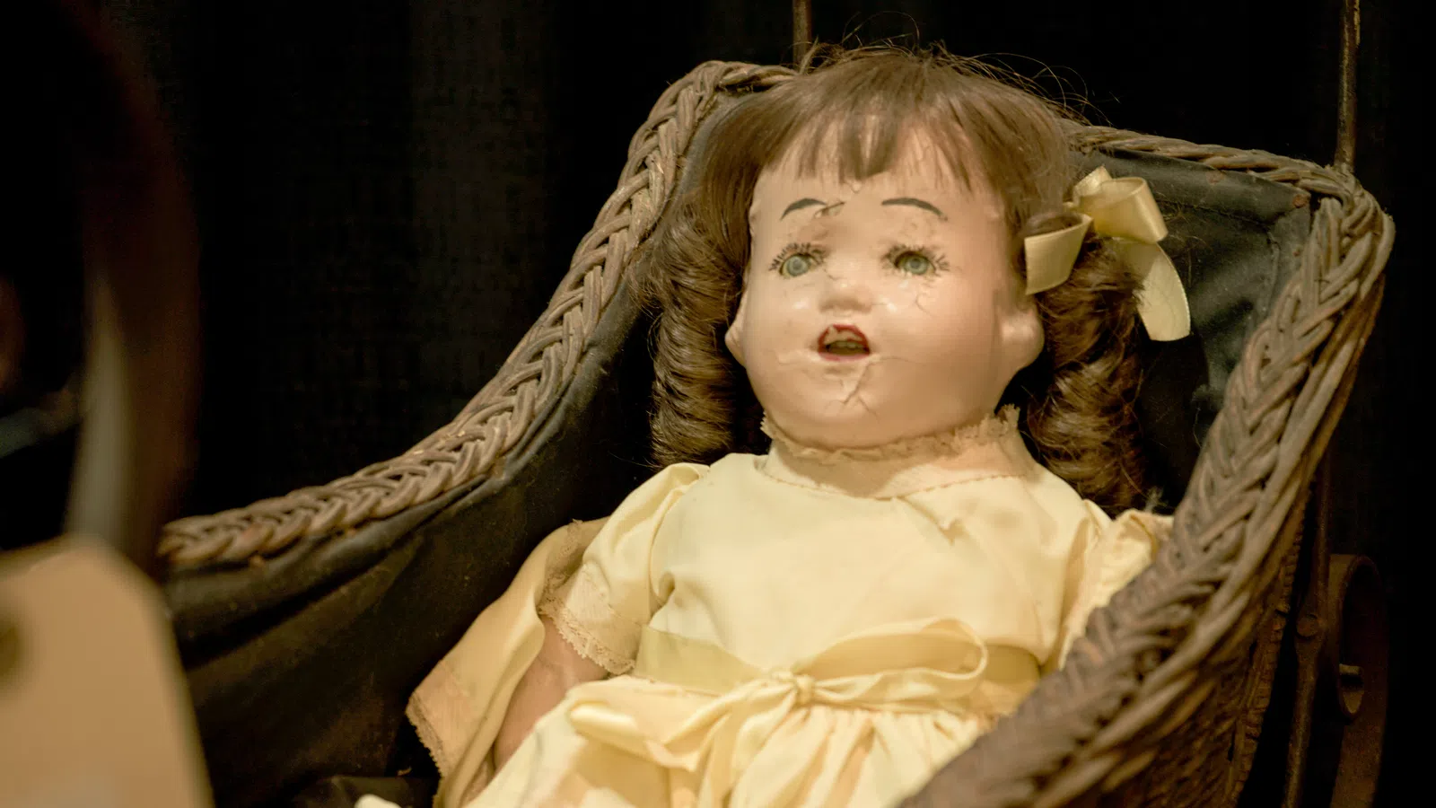 Vintage damaged doll with curly hair in a pale yellow dress sitting in a woven basket cradle.
