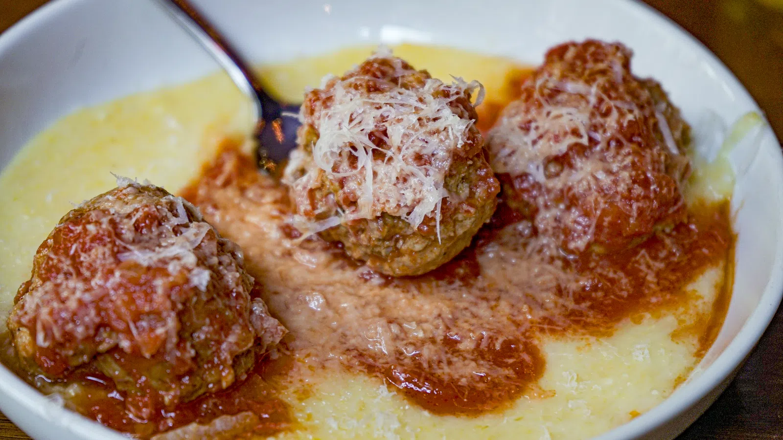Three meatballs topped with grated cheese served in marinara sauce over creamy polenta in a white bowl.