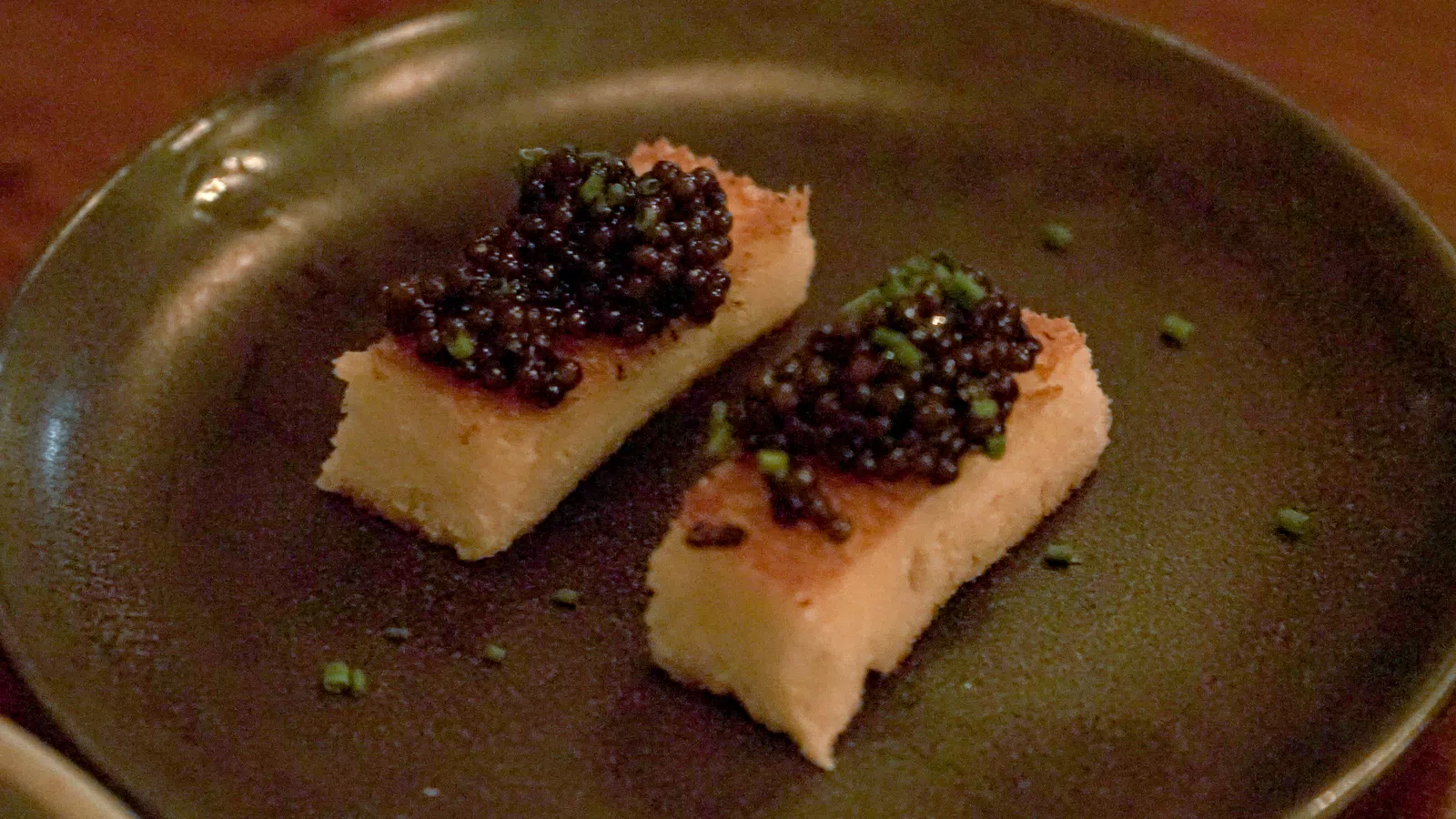 Two rectangular pieces of toasted bread topped with black caviar and garnished with chopped chives on a dark plate