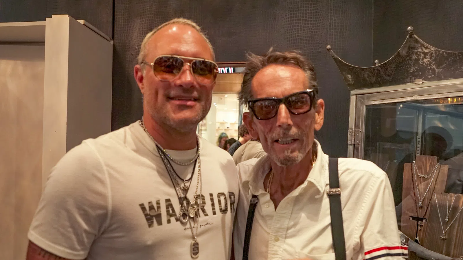 Two men wearing sunglasses and necklaces posing indoors at a jewelry store with display cases behind them