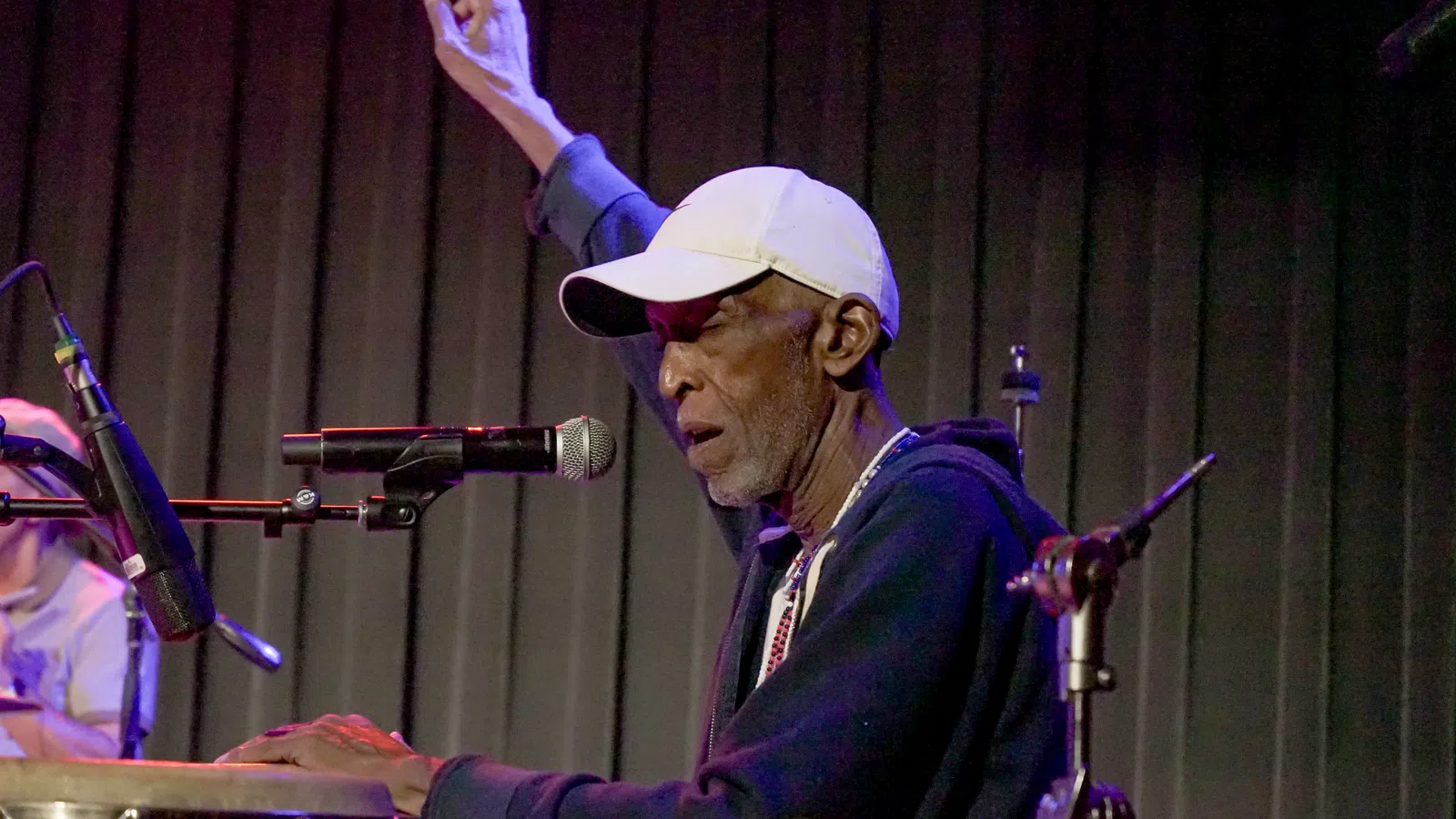 Elderly male musician playing congas and singing into a microphone with one hand raised on stage.