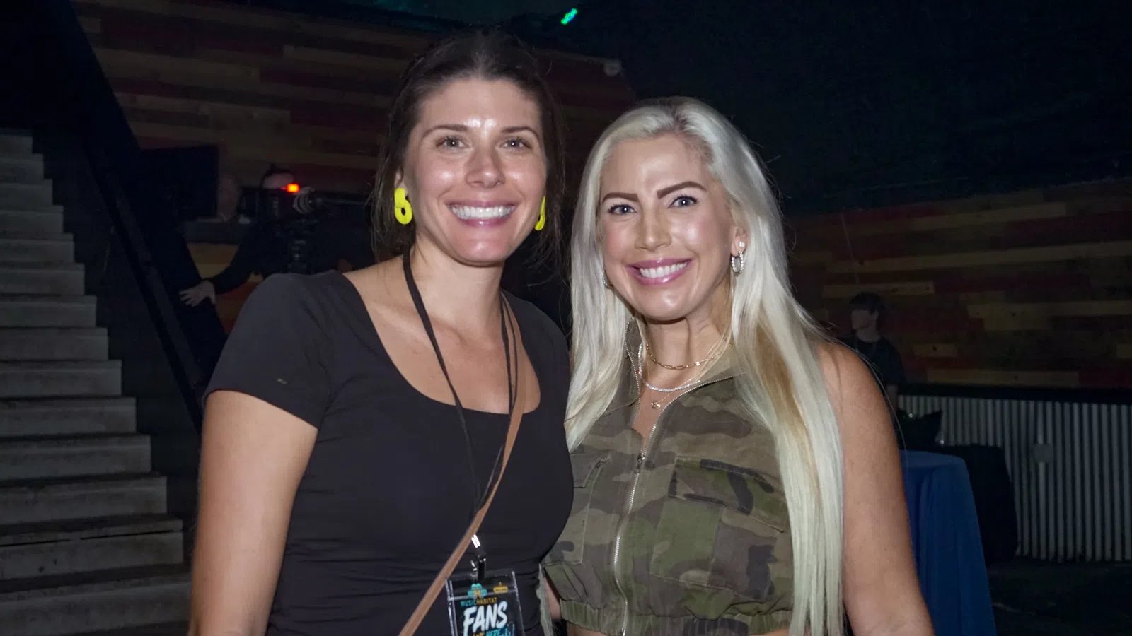 Two smiling women posing indoors, one in black top and the other in camouflage crop top with long blonde hair.