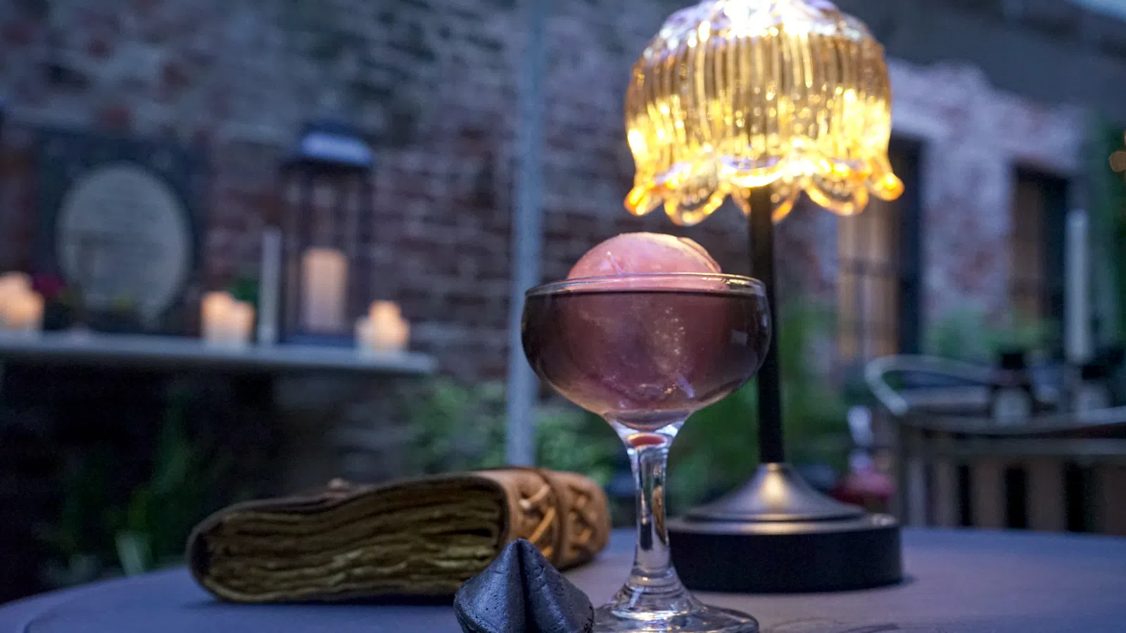 Elegant cocktail glass with a pink spherical garnish on a table beside a decorative lamp and fortune cookies.