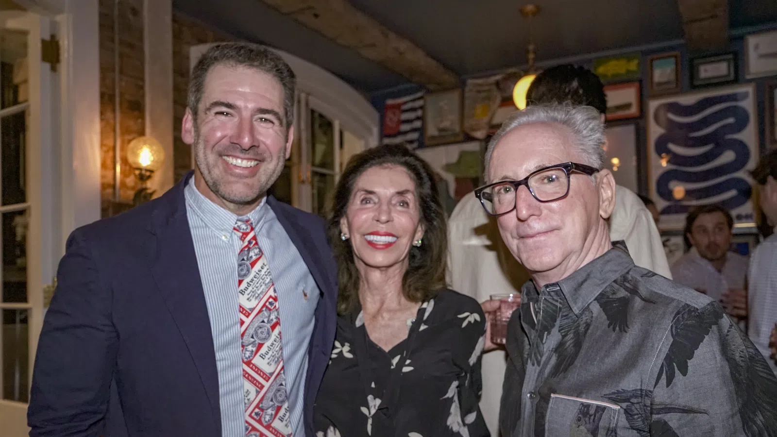 Three adults smiling together indoors, dressed in semi-formal attire at a social gathering with framed art on walls.