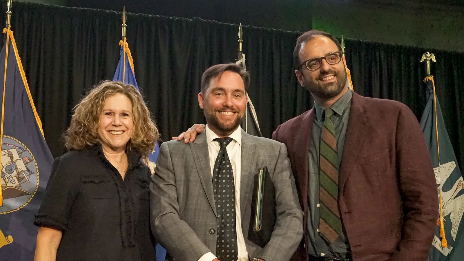 Three professionally dressed people smiling with flags in the background at an indoor event.