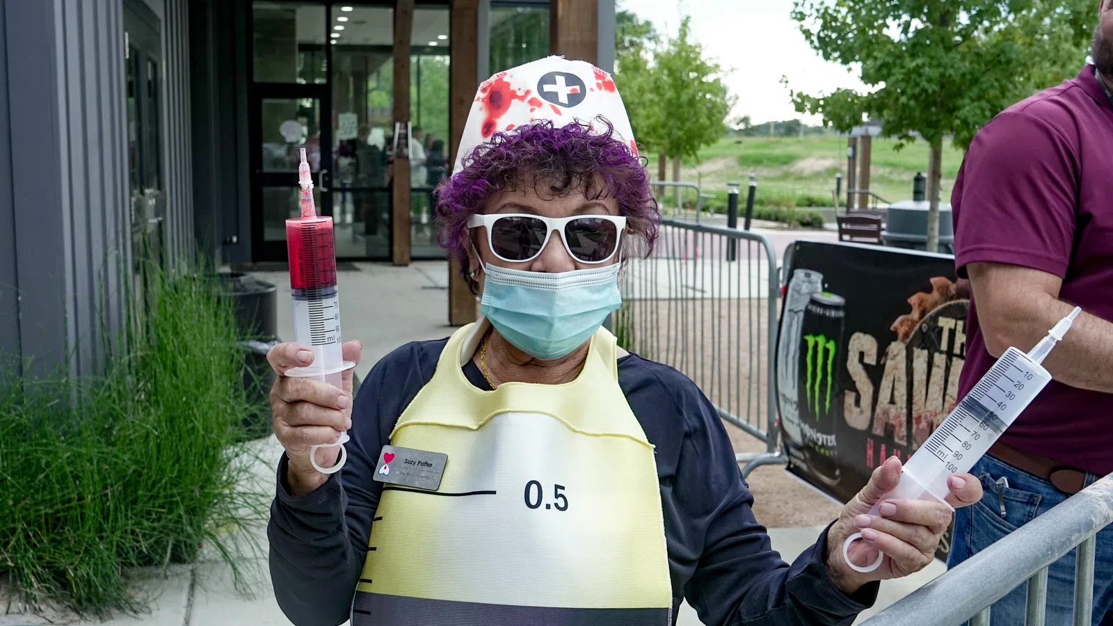 Person in nurse costume wearing sunglasses and mask holding two large syringes outside a building.