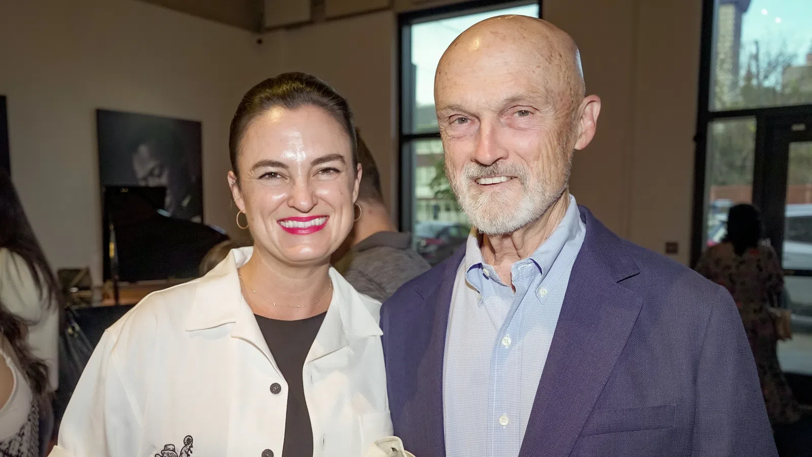 Smiling woman and elderly man standing indoors at a social event with large windows in the background.