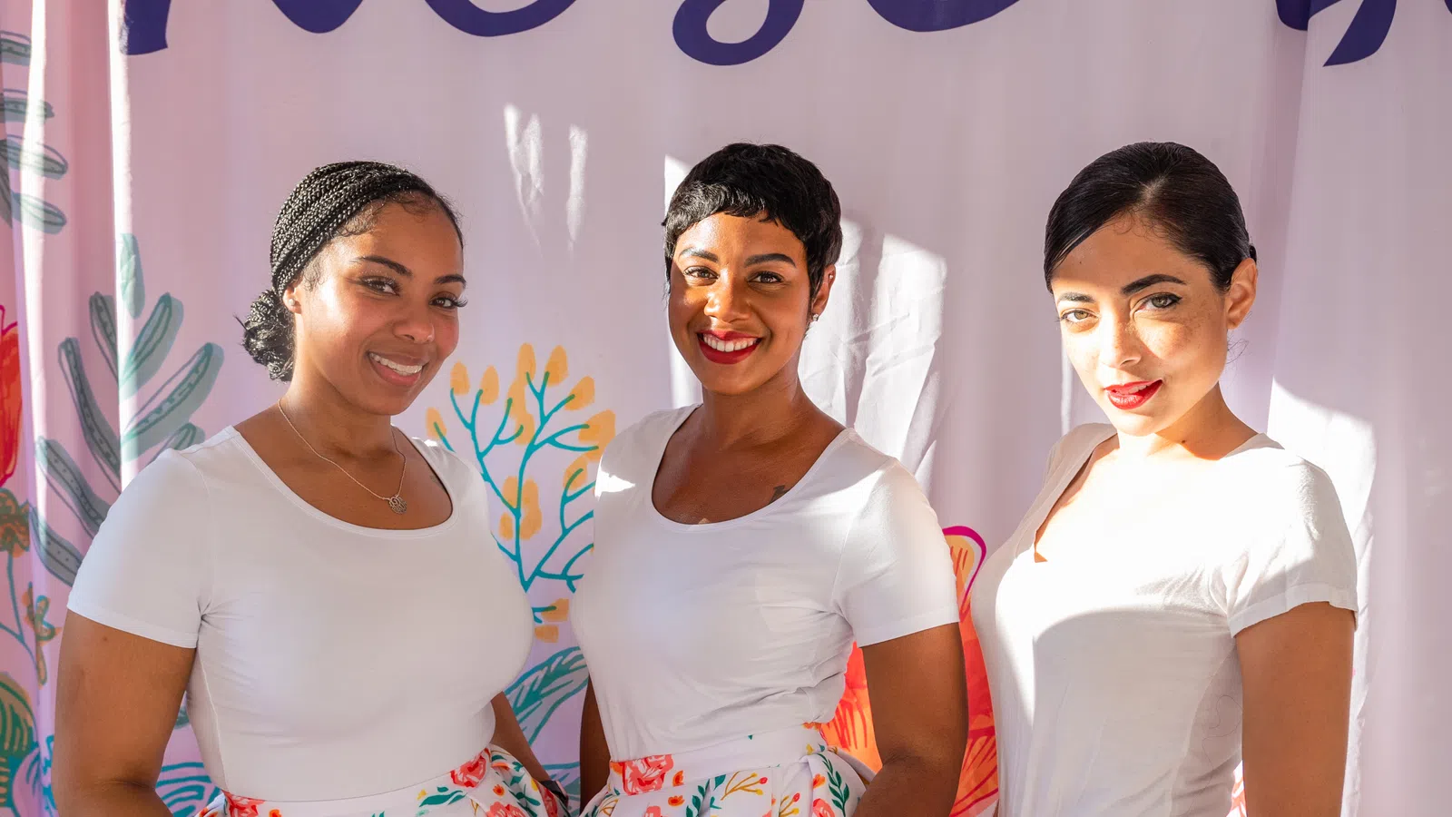 Three smiling women wearing white tops and floral skirts stand in front of a colorful backdrop with plants and coral designs.