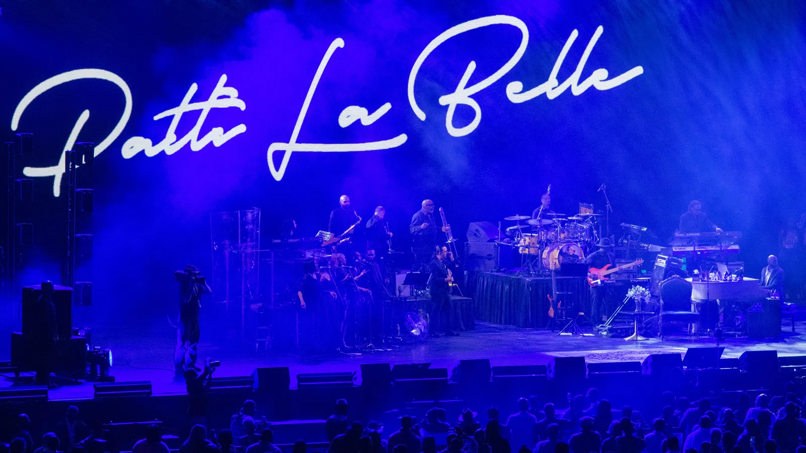 Patti LaBelle performing live on stage with band and blue lighting, audience visible in front of the stage