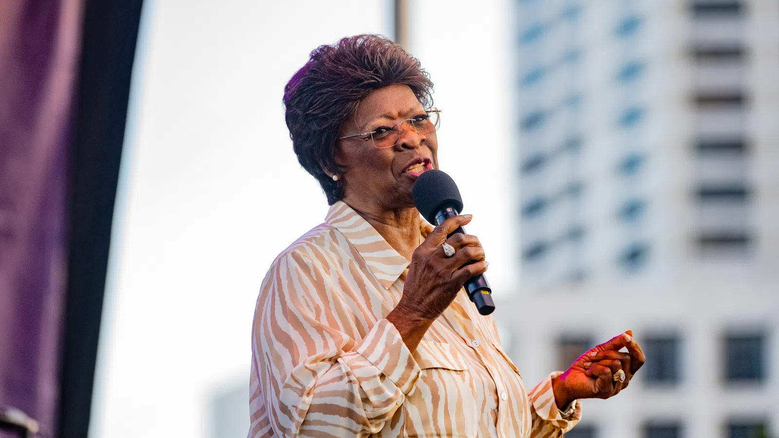 Elderly woman in striped blouse speaking into microphone at outdoor event with city building background