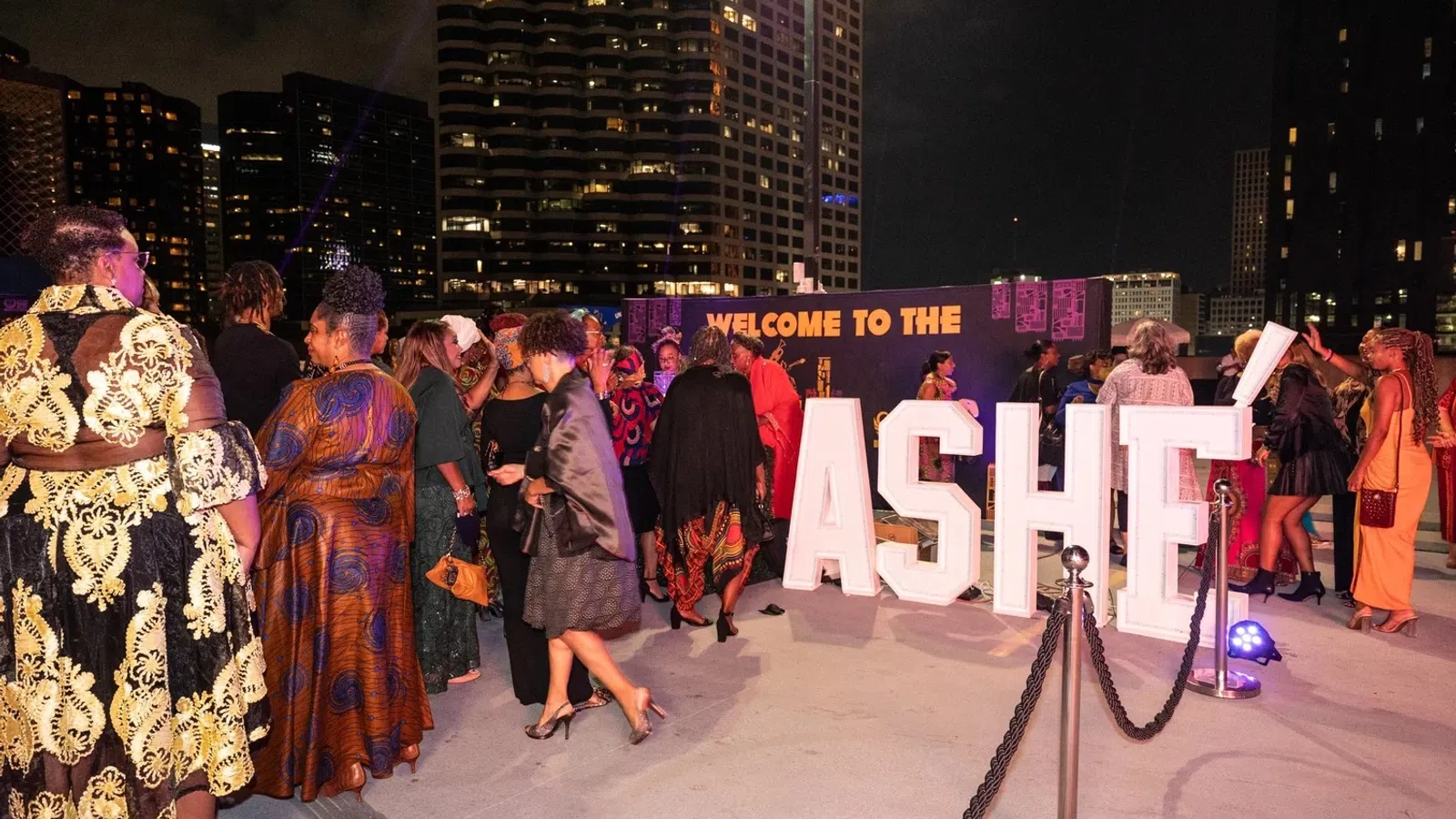 People dressed in elegant attire gather at a nighttime outdoor event with large illuminated ASHE letters and city buildings in the background