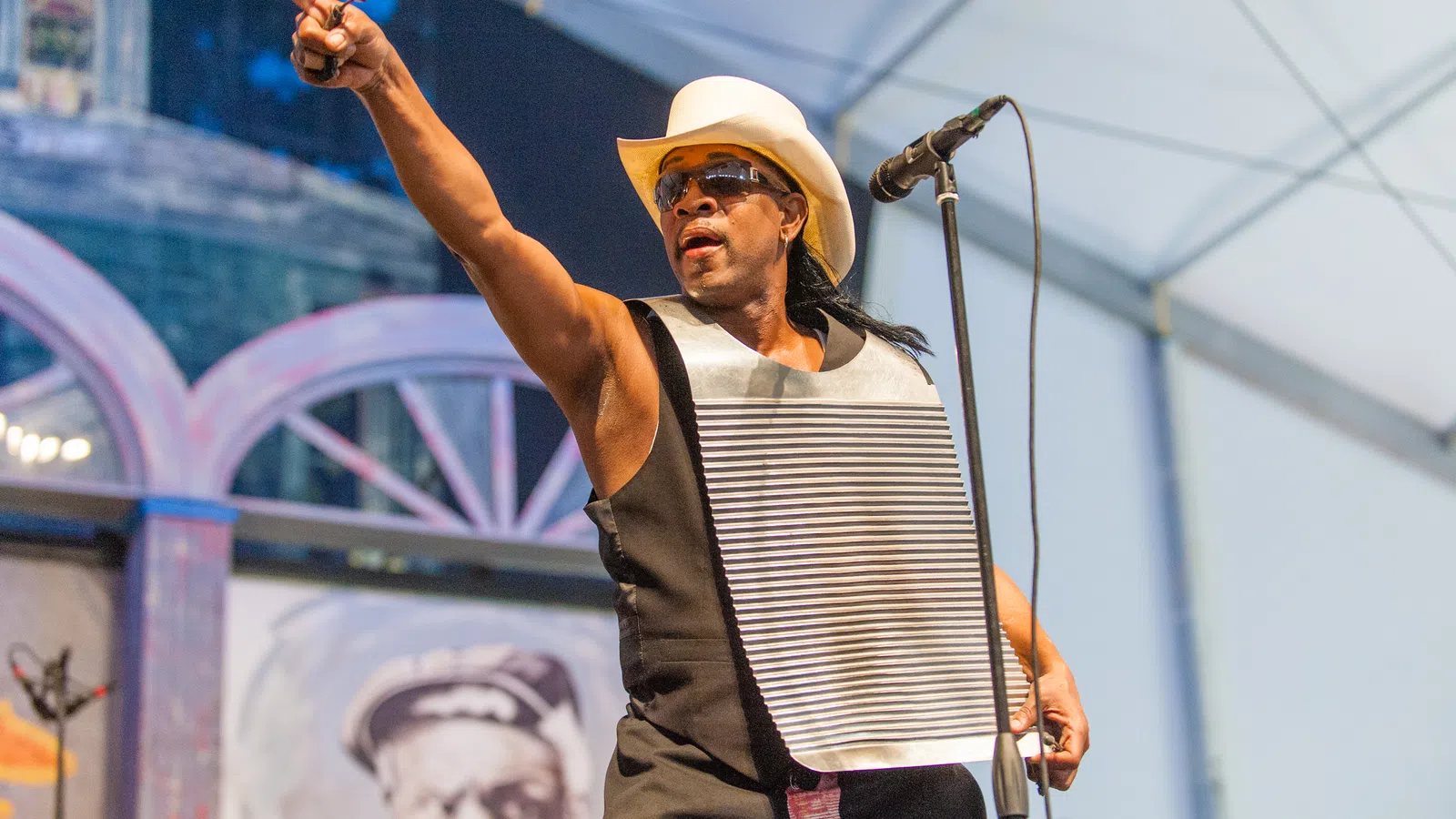 Musician wearing a white cowboy hat and playing a washboard while performing on stage with a microphone.