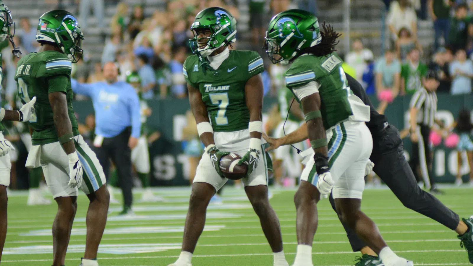Tulane football players in green uniforms huddle on field during game with crowd in background