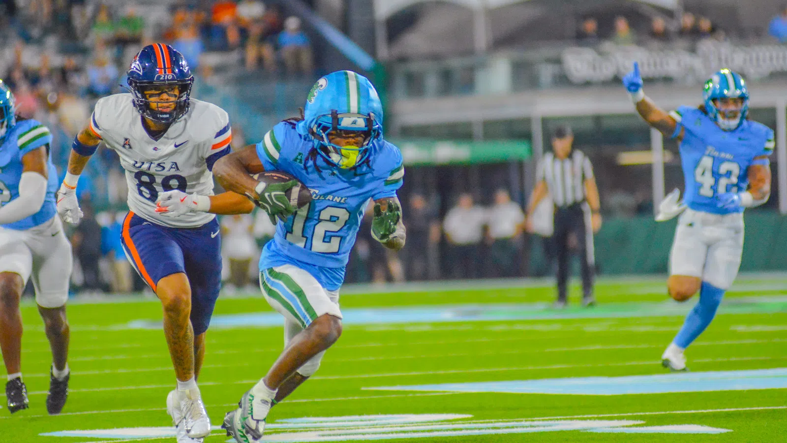 Football player in blue uniform running with ball as opponent in white uniform chases him on the field.