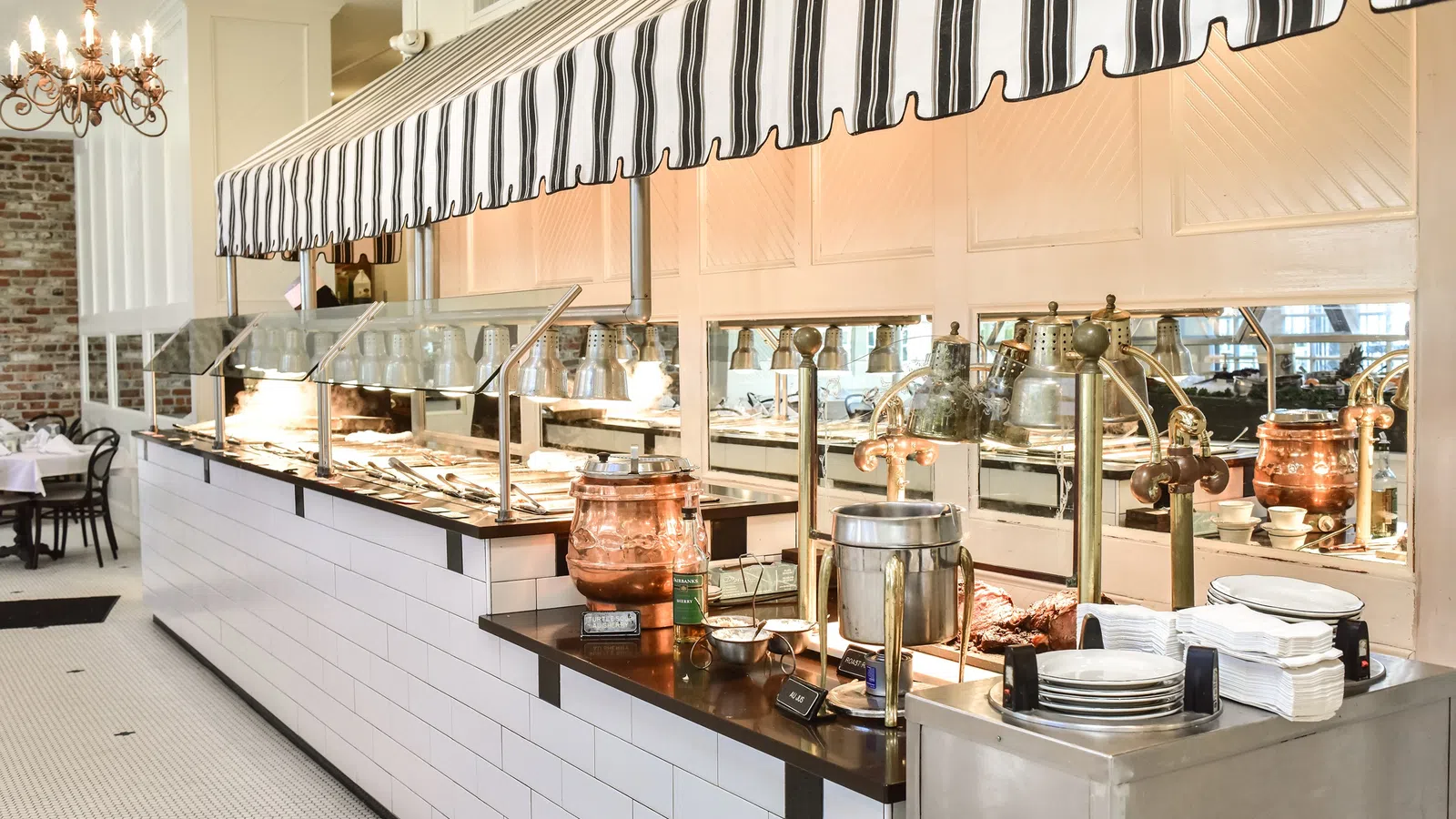 Bright buffet line with white tiles, striped canopy, copper pots, plates, and food warmers in a dining area.