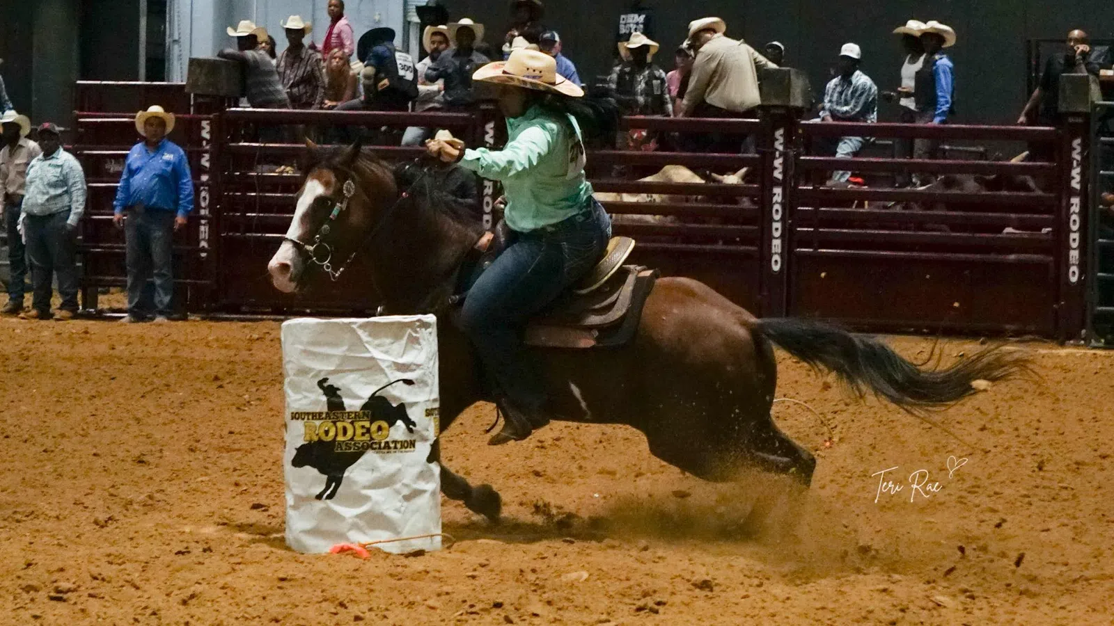 Cowgirl riding horse fast around barrel during rodeo competition in indoor arena with spectators