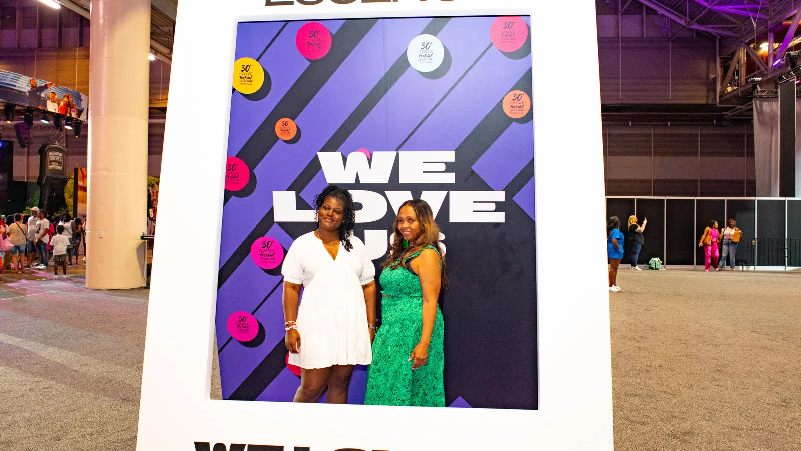 Two women posing inside a large Essence-themed photo frame with the text We Love Us at an indoor event.