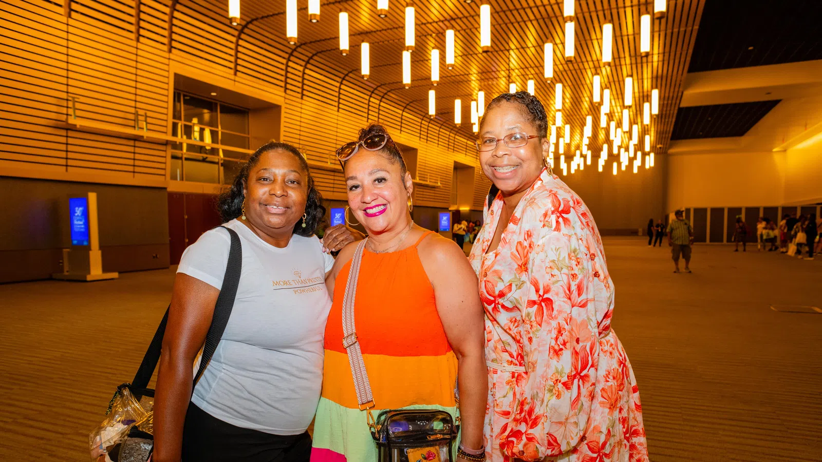 Three women smiling indoors under modern ceiling lights with wooden wall panels in a spacious venue.