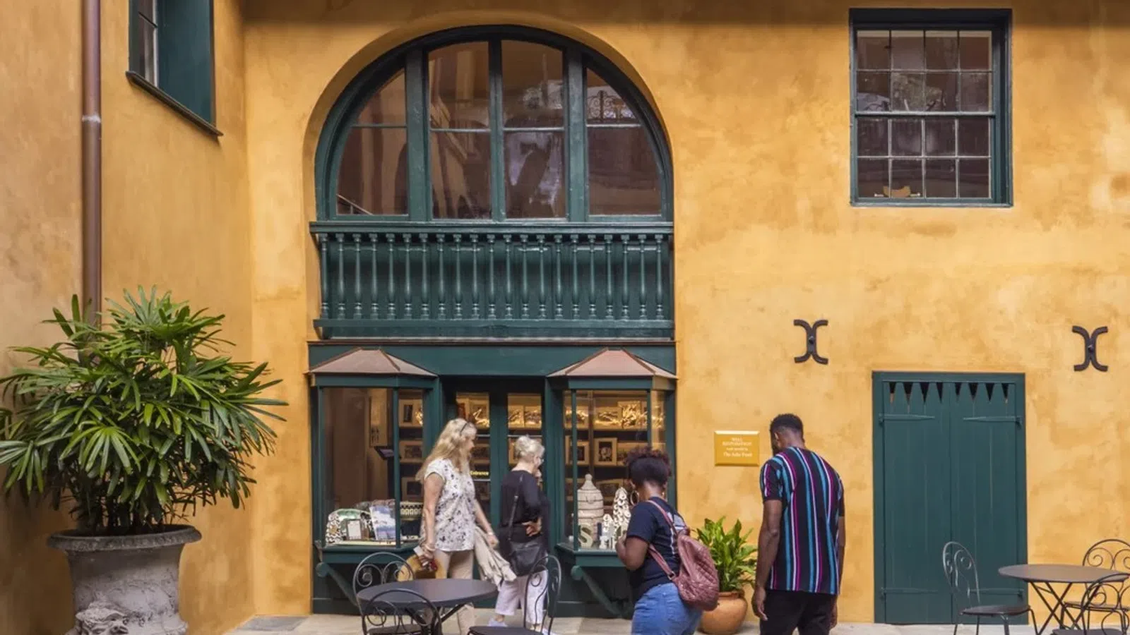 People walking and browsing outside a mustard-yellow building with green framed windows and outdoor tables.