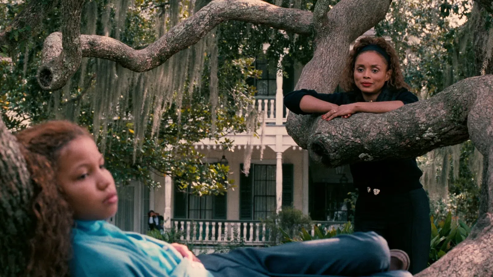 Two women relaxing on large tree branches with a white house and Spanish moss in the background.