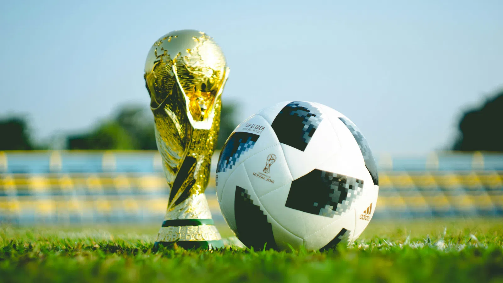 Golden FIFA World Cup trophy and official Adidas soccer ball on grass pitch with blurred stadium seats background.