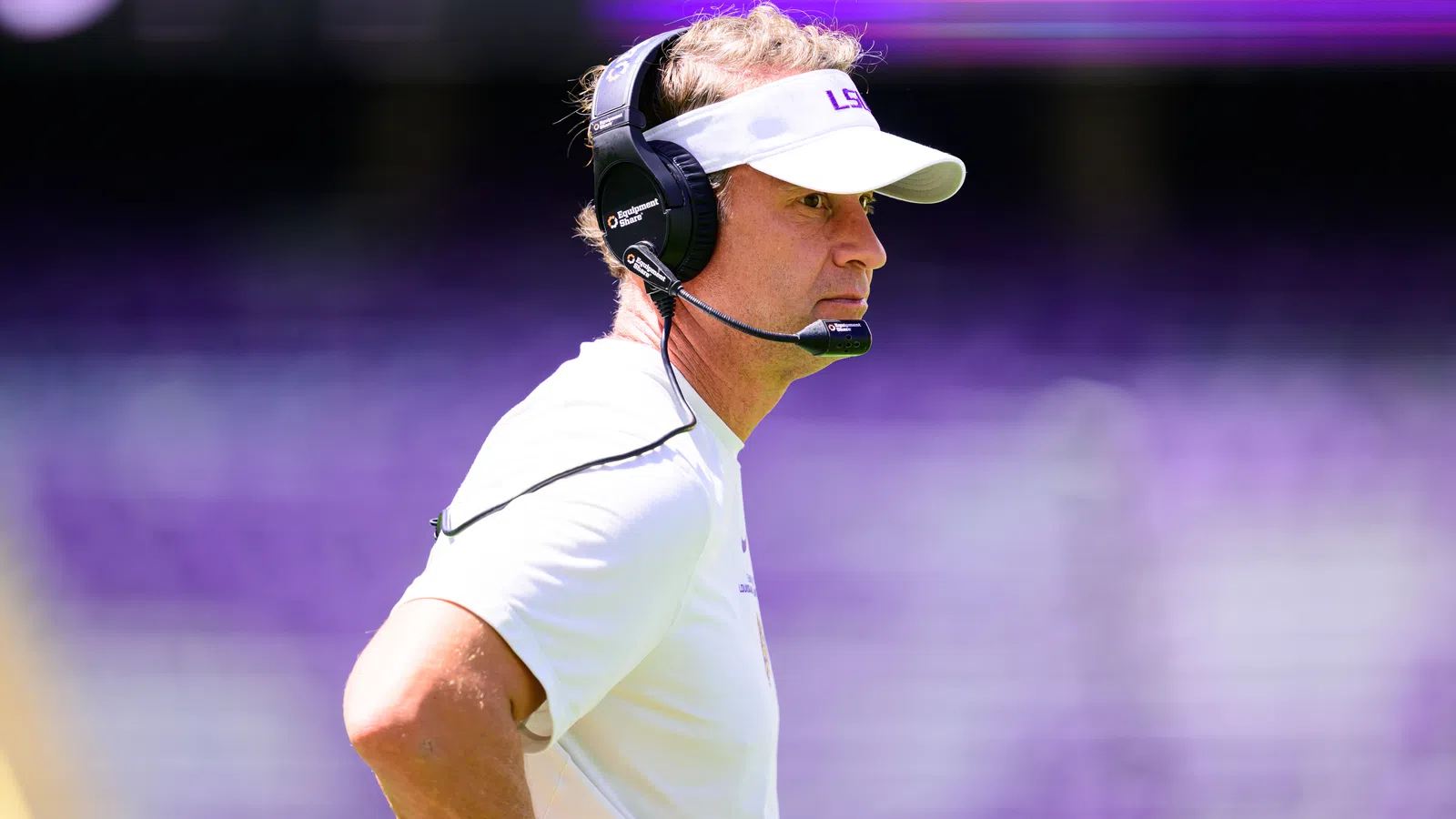 Football coach wearing LSU visor and headset focused during a game with blurred purple stadium background
