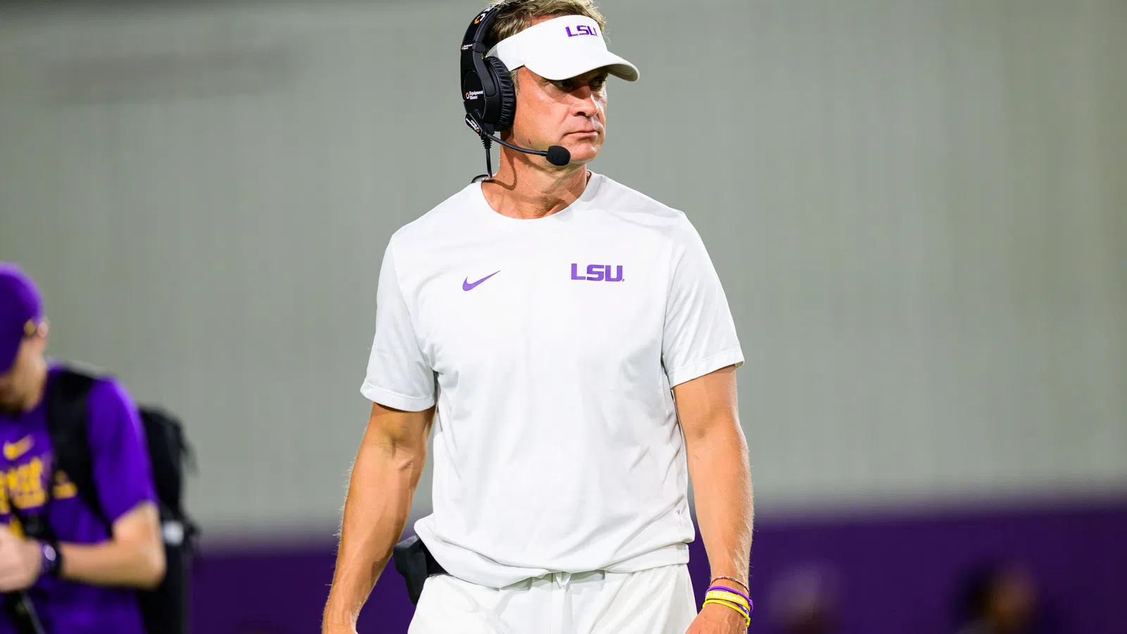 LSU football coach in white LSU gear and visor wears a headset on the sidelines during a game.