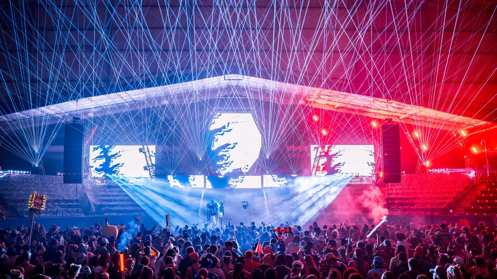 Crowd enjoying a nighttime concert with vibrant laser lights and colorful stage effects under a large canopy.