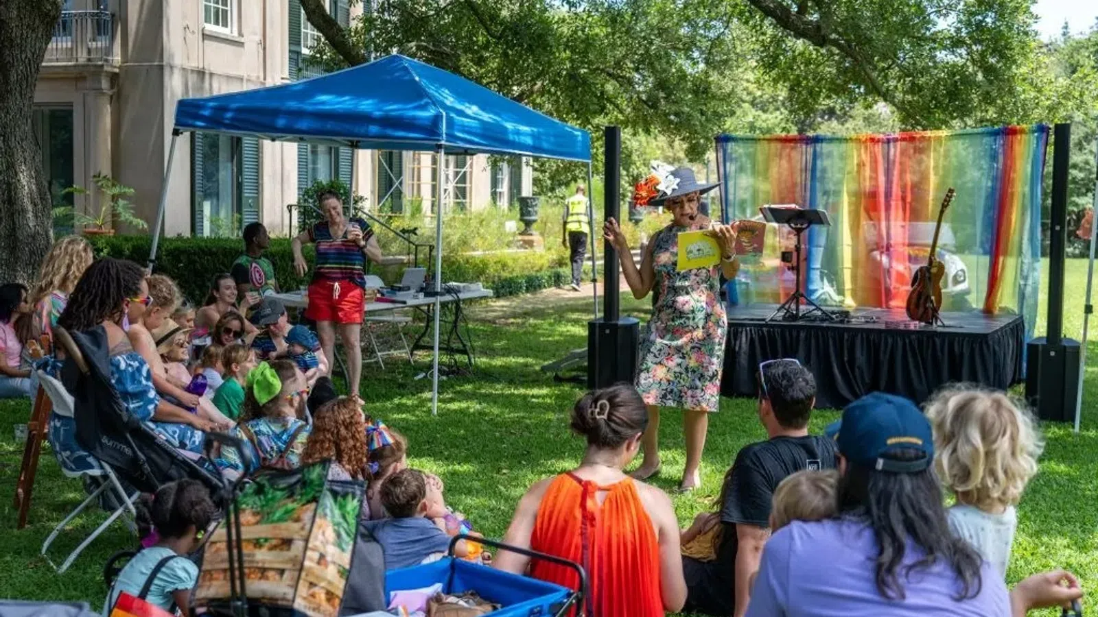 Woman reads a children's book outdoors to a seated diverse group of kids and adults on a sunny day