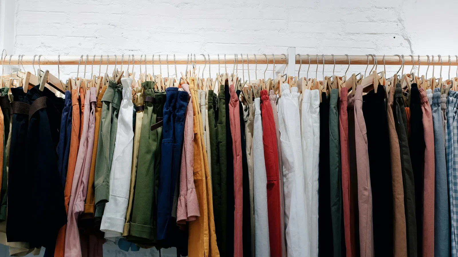 Variety of colorful pants and trousers hanging on a wooden rack against a white brick wall.