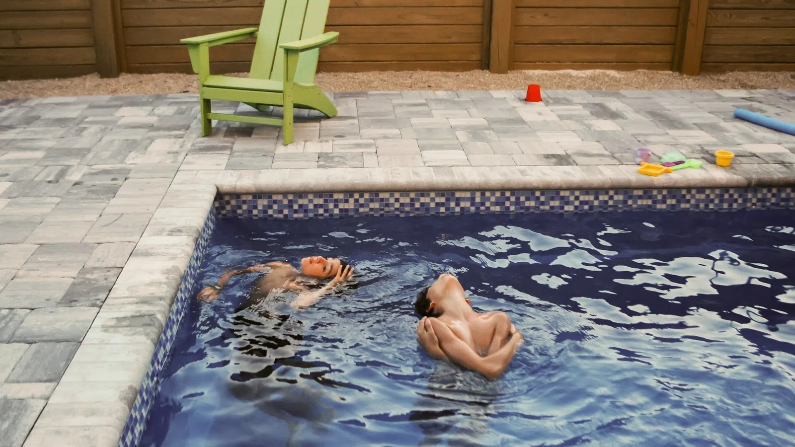 Two boys swimming and playing in a backyard pool next to a green Adirondack chair on a stone patio.