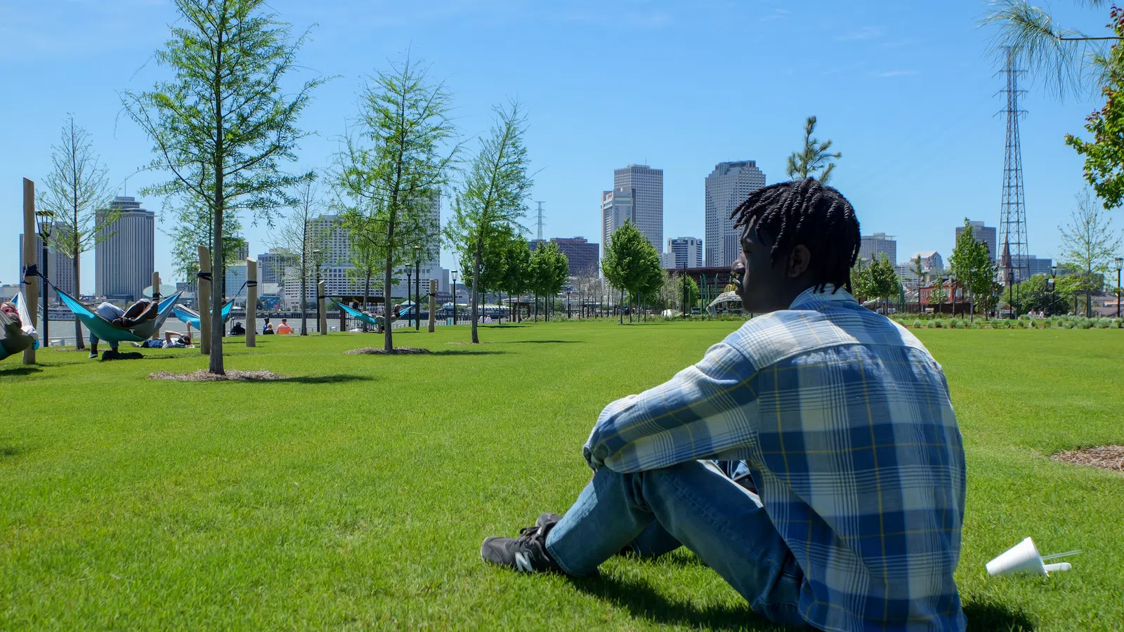 Young man in plaid shirt sitting on green grass with city skyline and trees in background on clear day