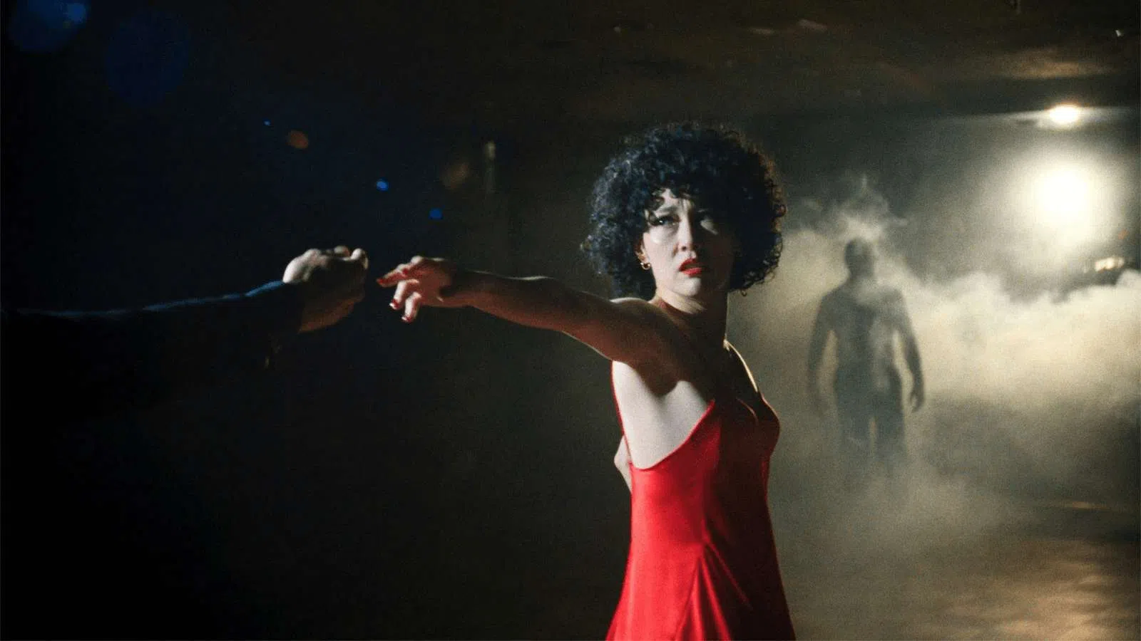 Woman in a red dress reaching out hand in a dark room with mist and a shadowy figure in the background