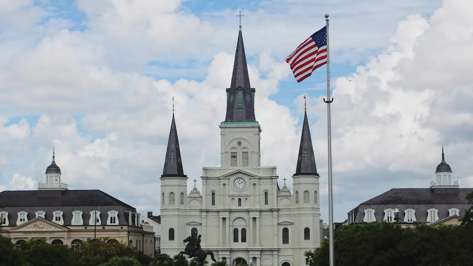 St. Louis Cathedral with three spires, American flag in foreground, cloudy sky backdrop in New Orleans