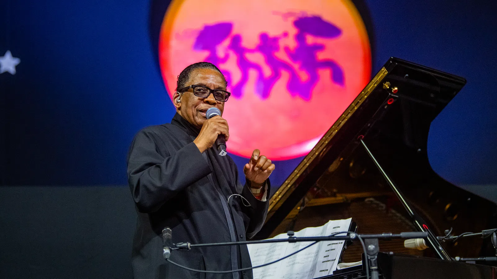 Jazz musician performing on stage with microphone and grand piano under colorful spotlight with dancing silhouettes.