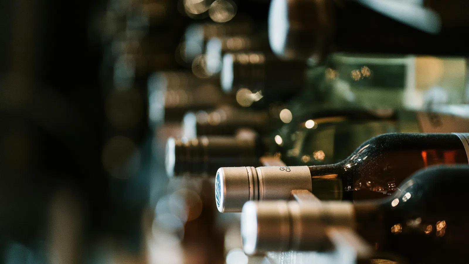 Close-up of wine bottles stacked horizontally on a wooden rack with dim lighting in a cellar or bar.