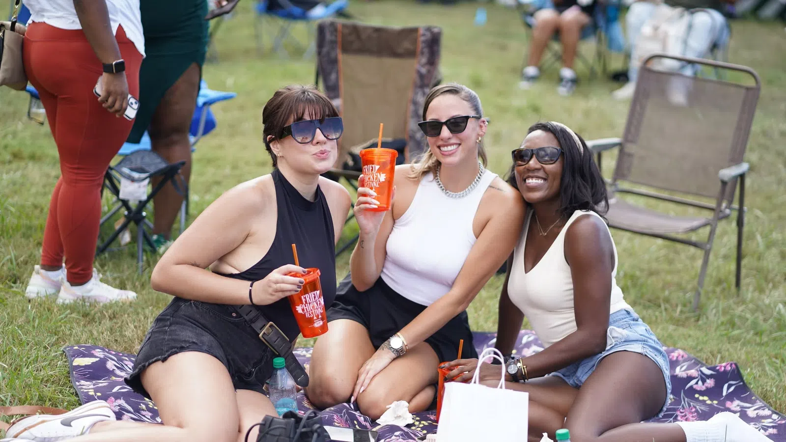 Three women wearing sunglasses enjoying drinks while sitting on a blanket at an outdoor event on grass.