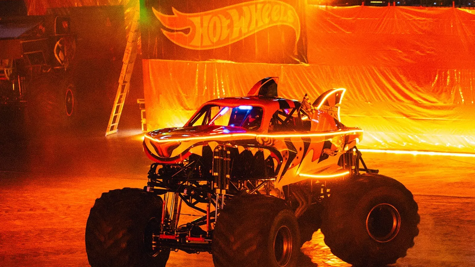 Monster truck with tiger stripes and glowing accents performs indoors in front of Hot Wheels banner at night.