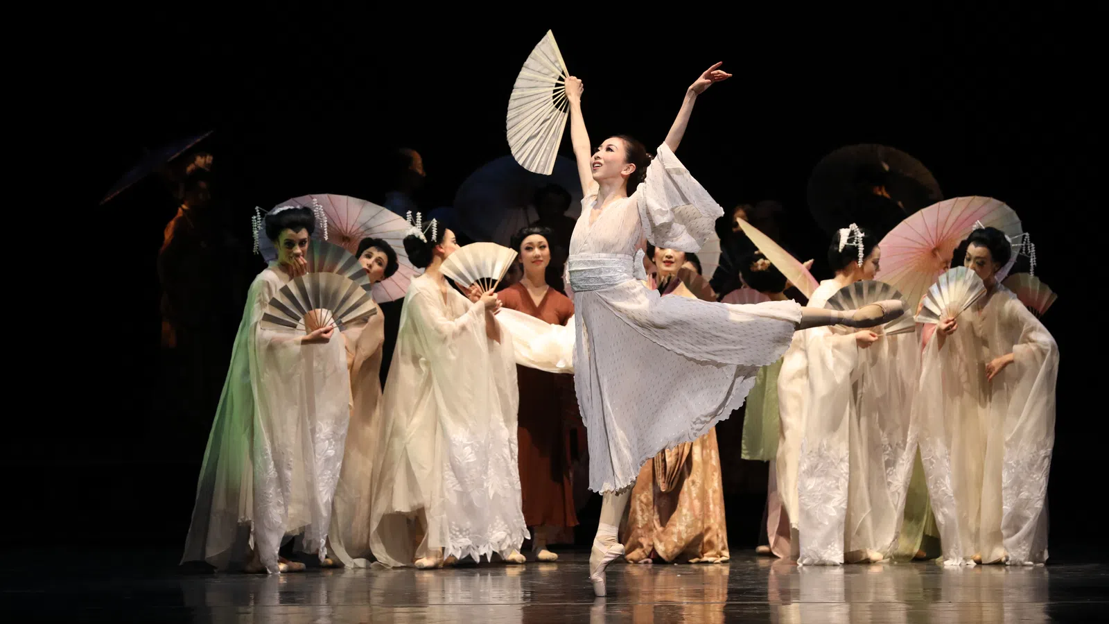 Ballet dancer in white costume performs with fans on stage with other dancers in traditional attire.