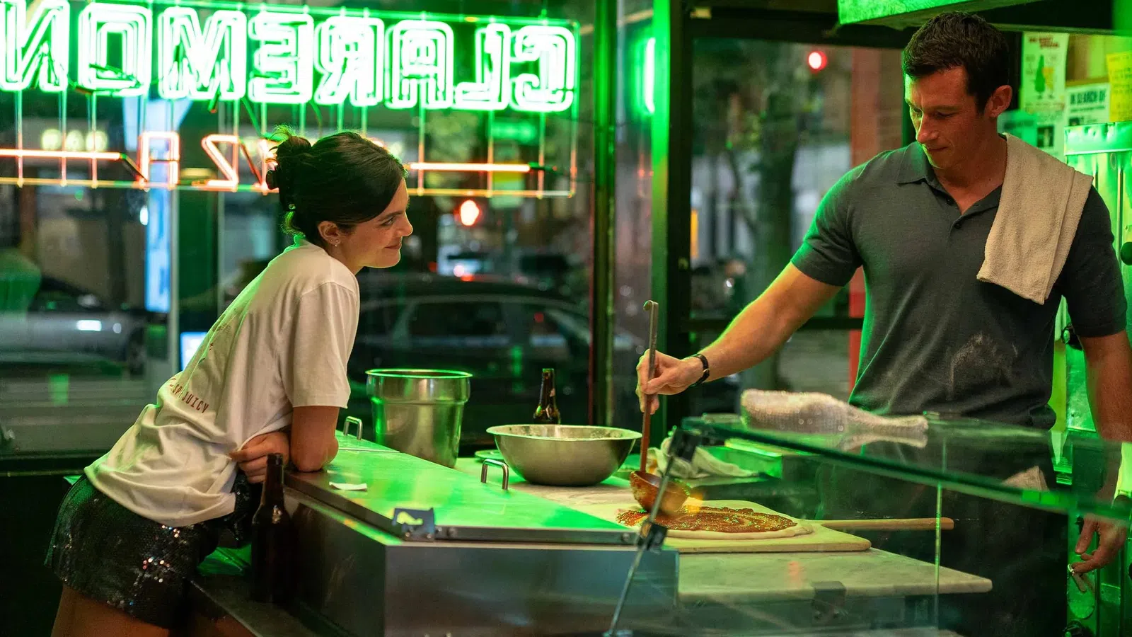 Man preparing pizza behind counter while woman watches inside a neon-lit pizzeria at night.
