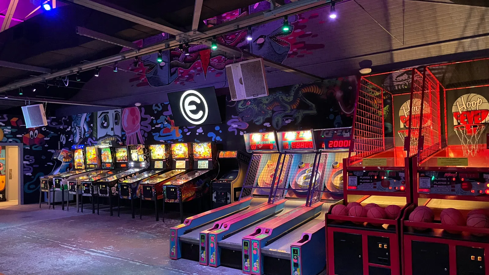 Indoor arcade with pinball machines, skee ball lanes, and basketball hoops under neon lighting.