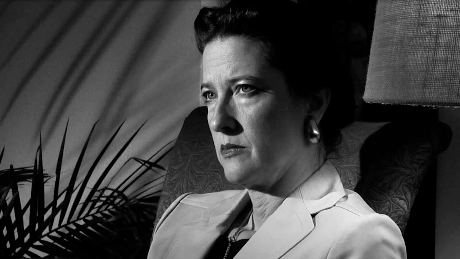 Black and white portrait of a serious woman sitting in a chair with shadows and a lamp in the background