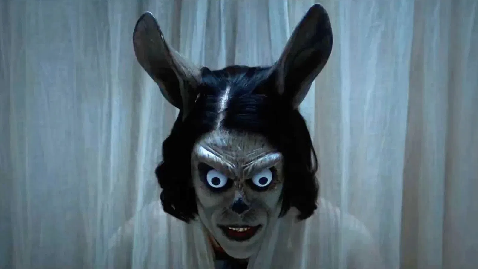 Creepy figure with exaggerated eyes, rabbit ears, and dark hair peers through white curtains in dim light.