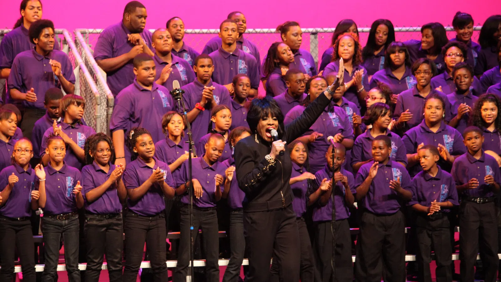 Choir of youths in purple shirts singing on stage with a soloist leading in black outfit and pink background.